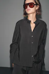 Camisa negra de lyocell con cuello redondo, manga larga con puños y bajo asimétrico con aberturas laterales. Cierre frontal con botones.