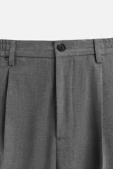 Pantalón gris de corte ancho con cintura elástica, pliegues frontales, bolsillos laterales y traseros de vivo. Incluye detalle de hebilla metálica en la trabilla y cierre frontal con cremallera y botón.