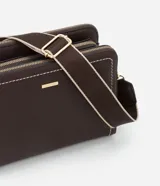 Cartera transversal de material sintético color marrón oscuro, de forma rectangular y estructura rígida. Presenta pespuntes decorativos en color blanco en los bordes frontales y cierres metálicos dorados. Incluye un asa ancha desmontable con diseño de rayas beige y marrón.