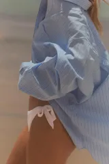 Camisa celeste con rayas verticales blancas, de corte amplio, cuello clásico, manga larga con puños abotonados, bolsillo de plastrón en el pecho y cierre frontal con botones.