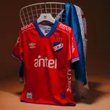 Camiseta de fútbol Umbro del Club Nacional de Football, modelo Away 1 2025. Diseño rojo con detalles en azul en cuello y mangas. Incluye logo de Umbro, escudo del club y sponsors en el frente.