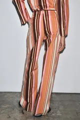 Pantalón de tiro medio con estampado de rayas verticales en tonos marrón, naranja, rosa y beige. Cuenta con trabillas en la cintura, cinturón ajustable con lazadas, bolsillos laterales y cierre frontal con cremallera y botones.