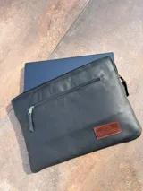 Funda negra de cuero para notebook, con cierre superior y bolsillo externo con cierre. Tiene un logo de la marca SierraMora en una etiqueta de cuero marrón cosida en la parte inferior derecha.