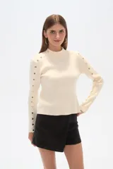 Sweater color crema de punto, con cuello alto y corte ajustado. Presenta detalle de tachas plateadas en hombros y mangas largas.