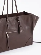 Bolso tote negro de cuero vacuno con textura granulada. Presenta cierres funcionales en los laterales para expandir el espacio y dos asas de mano. Incluye un tag colgante con aplique metálico y un tirador de cremallera con forma de figura.