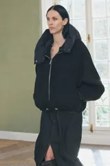 Chaqueta negra de cuello redondo confeccionada en hilatura con 50% lana. Tiene capucha extraíble, manga larga con puño interior, bolsillos delanteros y bajo ajustable con cordón elástico. Cierre frontal con cremallera metálica.