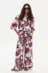 Pantalón ancho de lino con estampado floral grande en tonos morados sobre fondo blanco/crudo. Presenta cintura con elástico o ajuste y corte holgado en pierna.