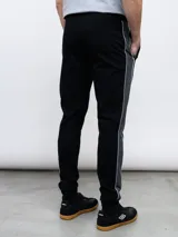 Pantalón deportivo Umbro UNY de piqué negro con puños ajustados, cintura elástica con cordón y logo Umbro estampado en blanco. Presenta una franja gris con ribete blanco a lo largo de la pierna. Confeccionado en poliéster y algodón.