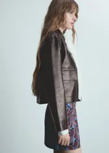 Chaqueta corta de poliuretano color marrón oscuro con textura, cuello camisero, manga larga con puños abotonados, dos bolsillos de solapa en la parte delantera y cierre frontal con botones.