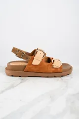 Sandalias marrones de cuero con suela plana y dos correas anchas con hebillas doradas.