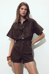 Mono corto color verde oliva, de denim con cuello solapa y manga corta. Tiene bolsillos delanteros y traseros, cintura elastizada en la espalda y cierre frontal con cremallera y botones metálicos.