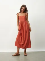 Vestido midi de color naranja, con escote recto y tirantes finos que se anudan sobre los hombros. Posee una cintura entallada con frunces que dan paso a una falda fluida.
