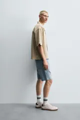 Bermuda de denim de algodón, color gris lavado, con corte relaxed fit y roturas visibles en la parte delantera de ambas piernas. Presenta cinco bolsillos y cierre frontal con cremallera y botón.