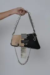 Cartera bandolera de cuero sintético color negro, con diseño patchwork y detalles de tachas y hebillas metálicas. Tiene asa de cadena plateada y cierre con solapa y broche.