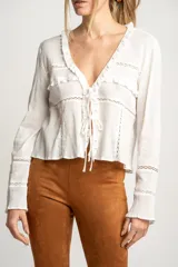 Blusa blanca de algodón con escote en V, cierre con lazos, mangas largas con puños elásticos y detalles de encaje y volados.