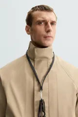 Parka ligera color beige, de corte relaxed fit, confeccionada en tejido técnico repelente al agua. Tiene cuello alto, mangas largas con puños ajustables con botón a presión, bolsillos de solapa en la cadera y cierre frontal con cremallera oculta por solapa con botones a presión.