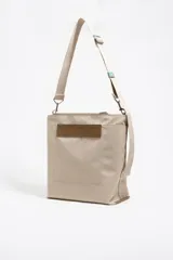 Bolso tipo bucket de nylon color beige, con estructura blanda, cierre de cordón y asa de tejido regulable. Presenta logo metálico de Bimba y Lola en el frente.