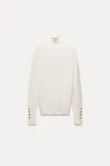 Jersey negro de punto liso con cuello alto, manga larga y detalle de botones dorados en los puños.