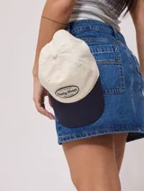 Gorra de lona color beige con visera azul marino y parche bordado con el logo de Rusty Shop.