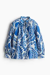 Blusa oversized con cuello elevado fruncido, abertura en V adelante y bajo recto. Mangas globo largas de corte raglán con abertura y botón en los puños. Estampado floral en tonos azules y blancos.