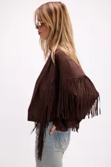 Chaqueta de efecto gamuza en color tostado, con solapas amplias, cierre frontal con un botón y flecos largos decorativos en la parte frontal y trasera de las mangas. Presenta una estructura definida y forro interior.