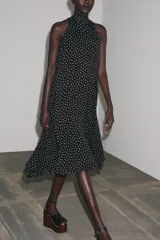 Vestido midi negro con estampado de lunares blancos, cuello halter con pañuelo integrado, sin mangas y bajo asimétrico. Cierre con abertura y botón en la nuca.