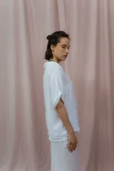 Camisa color crudo confeccionada en lino, de corte holgado, con cuello clásico, mangas cortas con dobladillo y cierre frontal con botones. Presenta detalle de pliegues verticales en la parte frontal.