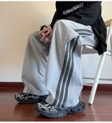 Pantalón deportivo gris claro, estilo cargo o jogging, con franjas negras verticales en los laterales de las piernas. Posee tobillos regulables y cintura ajustable.