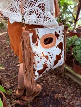 Bandolera de cuero con estampado animal print de vaca en tonos marrón y blanco, con flecos de cuero marrón en los laterales. Tiene correa larga de cuero marrón regulable y asa de mano con forma ovalada.