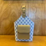 Bolso matero con diseño a cuadros color lila y blanco, con bolsillo frontal color beige y manija superior con hebilla.