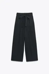 Pantalón negro con finas rayas verticales blancas, de tiro alto y corte ancho. Incluye cinturón de lazo en el mismo tejido, bolsillos laterales y pinzas.