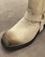 Bota de cuero color hueso, con caña media, punta cuadrada y suela track color negro. Presenta una correa con hebilla metálica alrededor del tobillo, que puede ser removida.