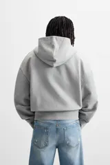 Sudadera gris con capucha, corte holgado, cierre frontal con cremallera, bolsillos laterales ocultos y terminaciones irregulares.
