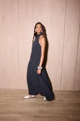 Vestido largo negro, holgado, con escote halter y espalda baja. Presenta un detalle de volado en el cuello y un maxi volado en la cadera. El material es tela Cupro, con apariencia similar a la gamuza.