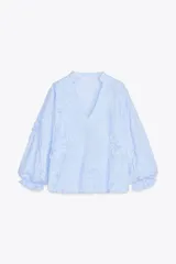 Blusa celeste con rayas verticales blancas, cuello redondo con escote en pico y mangas largas abullonadas terminadas en puño elástico. Presenta detalle de tejido en relieve con forma de flor.