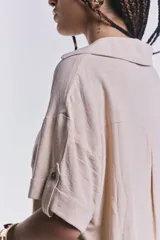 Camisa color beige de corte cropped, con cuello clásico, cierre frontal con botones y dos bolsillos de parche en el pecho.