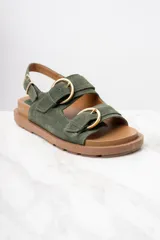 Sandalias de gamuza color verde militar con dos tiras anchas sobre el empeine y una tira trasera, todas ajustables con hebillas doradas. Suela plana color marrón.