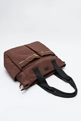 Bolso tote marrón de nylon con doble asa de mano negra y correa bandolera negra regulable y desmontable. Tiene dos bolsillos frontales con cierre metálico dorado.