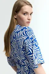 Vestido midi camisero con estampado de hojas blancas sobre fondo azul. Tiene cuello camisero, botonadura frontal, mangas cortas raglán, elástico en la cintura y cinturón extraíble para atar.