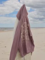 Buzo con capucha color lila, de algodón y fit relajado. Presenta un estampado con una palmera y el texto "Rainbow Bay" en el pecho.