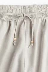 Short color beige de tiro alto con cintura elástica fruncida con ribete de volado, cordón para ajustar fino con cuentas de metal y bolsillos laterales discretos.