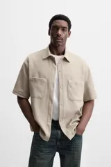 Sobrecamisa color beige de tejido elástico, con cuello solapa, manga corta y bolsillos de parche en el pecho. Cierre frontal con botones a presión.