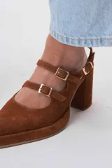 Zapato de cuero marrón con taco bloque, punta fina y dos tiras con hebillas doradas.