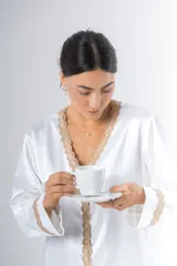 Pijama blanco de satén elastizado, compuesto por una camisa de manga larga con escote en V, abotonada adelante y con puntilla en escote y puños, y un pantalón recto con bolsillos laterales, elástico en la cintura y puntilla en el ruedo.