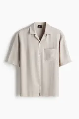 Camisa de manga corta color beige con cuello abierto, tapeta lisa, bolsillo superior y bajo recto. Corte estándar para una silueta clásica y cómoda.