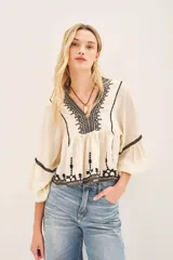Blusa color crema con bordados negros, escote en V y mangas largas abullonadas con terminaciones elásticas.