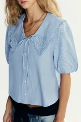 Camisa celeste a rayas verticales blancas, de corte recto, con cuello bobo con bordados y escote pico con lazada. Tiene mangas cortas abullonadas con elástico y cierre frontal con botones.