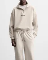 Conjunto deportivo color beige compuesto por un buzo con cierre de cremallera en el cuello y un pantalón de jogging con cintura elástica ajustable con cordón. Ambos con logo bordado en negro.