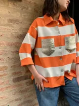 Camisa de manga 3/4 con cuello clásico y cierre frontal con botones. Diseño a rayas horizontales color naranja y blanco, con bolsillos frontales con solapa en color dorado.