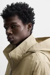 Parka color beige de tejido técnico ligero, corte relaxed fit, cuello alto con capucha plegable, manga larga con puños ajustables con botón a presión, cierre frontal con cremallera, cintura ajustable con elástico interno y múltiples bolsillos frontales de parche con solapa.
